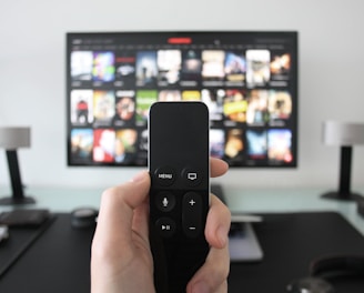 A person holds a remote control in front of a TV displaying a streaming platform interface with various movie and show thumbnails. The room includes a desk with two small lamps on either side of the TV.