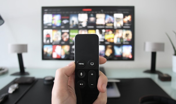 A person holds a remote control in front of a TV displaying a streaming platform interface with various movie and show thumbnails. The room includes a desk with two small lamps on either side of the TV.