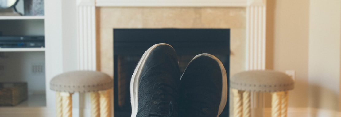 A person is relaxing in a living room with feet up on a wooden table. A TV mounted above a fireplace is displaying a popular streaming platform. Shelves on either side of the fireplace hold decorative items. A smartphone can be seen on the table.