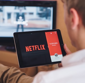 A person is sitting on a couch holding a tablet that displays the Netflix logo prominently. In the background, a television is showing a blurred image. The person appears to be engaged in using both devices simultaneously.