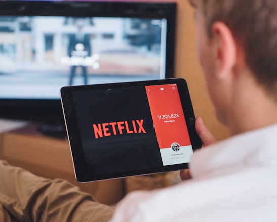 A person is sitting on a couch holding a tablet that displays the Netflix logo prominently. In the background, a television is showing a blurred image. The person appears to be engaged in using both devices simultaneously.