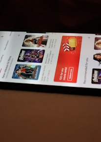 A smartphone displaying a streaming app interface, showing various movie and TV show thumbnails. The screen includes categories like 'For you' and 'New release hit films', with images from popular films. The background is a dimly lit setting with a fabric surface.