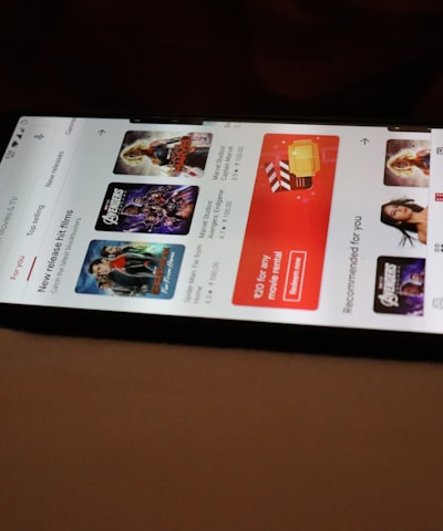 A smartphone displaying a streaming app interface, showing various movie and TV show thumbnails. The screen includes categories like 'For you' and 'New release hit films', with images from popular films. The background is a dimly lit setting with a fabric surface.