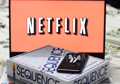 A screen displaying the Netflix logo in bold, black and white lettering against a vibrant red background. In front of the screen, there is a closed box for the board game 'Sequence', with a smartphone and wireless earbuds resting on top.