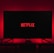 A dark room featuring a television screen displaying the word 'Netflix' in red against a black background. The screen is centered on a console table, with a video game controller placed in front. The room is dimly lit by red ambient lighting, creating a cozy atmosphere.