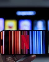 A hand holding a smartphone displaying the Netflix logo on its screen with a background of colorful vertical lines. In the background, a television screen shows blurry icons or profiles with various colors.