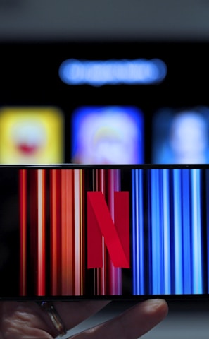 A hand holding a smartphone displaying the Netflix logo on its screen with a background of colorful vertical lines. In the background, a television screen shows blurry icons or profiles with various colors.