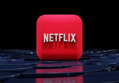 A red, cube-shaped icon with the word 'NETFLIX' prominently displayed in white, bold letters. The cube is set against a dark, glossy, circular patterned background, reflecting a sleek, futuristic design.