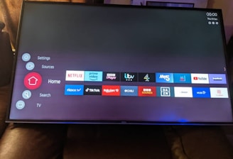 A television screen displays a smart TV interface with multiple app icons arranged in rows. Options such as Settings, Sources, Home, Search, and TV are listed on the left side. Some recognizable app icons include Netflix, Prime Video, BBC iPlayer, ITV Hub, All 4, My5, YouTube, and TikTok. The time and date are shown in the top right corner.