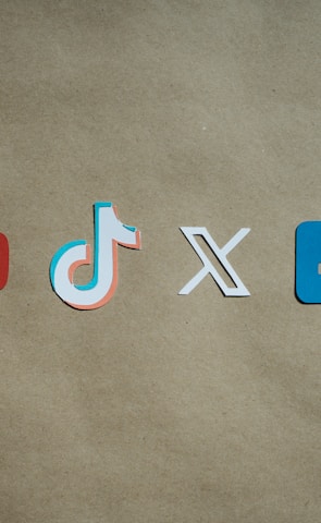 Four social media platform logos are arranged on a brown background. From left to right, the logos are for YouTube in red, TikTok in a combination of red, blue, and white, X in white, and Facebook in blue.