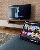 A modern living room features a flat-screen television mounted on a light-colored wooden stand with a patterned wall backdrop. Below the TV, a carpet lies on a wooden floor. In the foreground, a tablet displays a streaming platform interface with various show thumbnails, suggesting a relaxed entertainment setup.