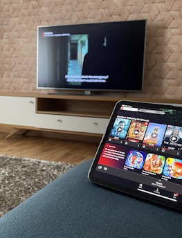 A modern living room features a flat-screen television mounted on a light-colored wooden stand with a patterned wall backdrop. Below the TV, a carpet lies on a wooden floor. In the foreground, a tablet displays a streaming platform interface with various show thumbnails, suggesting a relaxed entertainment setup.