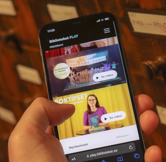 A person holding a smartphone displaying a webpage with a Scandinavian library streaming service. The screen shows two video thumbnails, one with a TV displaying a program and the other with a woman holding a book. The background features a wooden drawer set with labels.