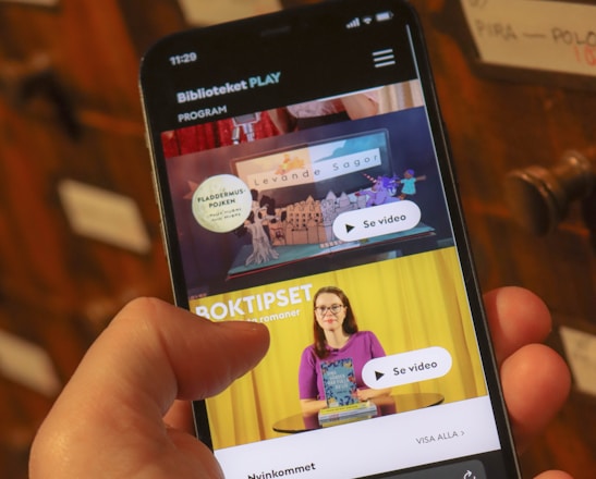 A person holding a smartphone displaying a webpage with a Scandinavian library streaming service. The screen shows two video thumbnails, one with a TV displaying a program and the other with a woman holding a book. The background features a wooden drawer set with labels.
